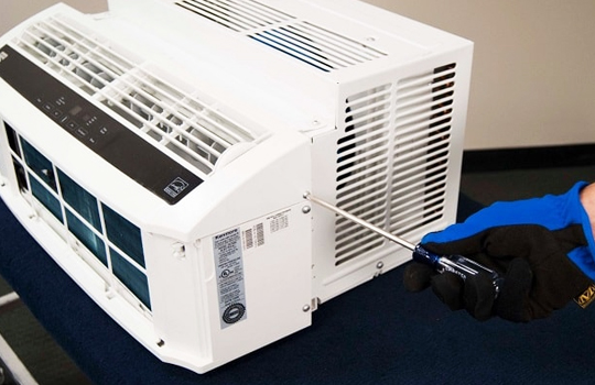 The technician is wearing gloves and servicing a white window AC installation unit.